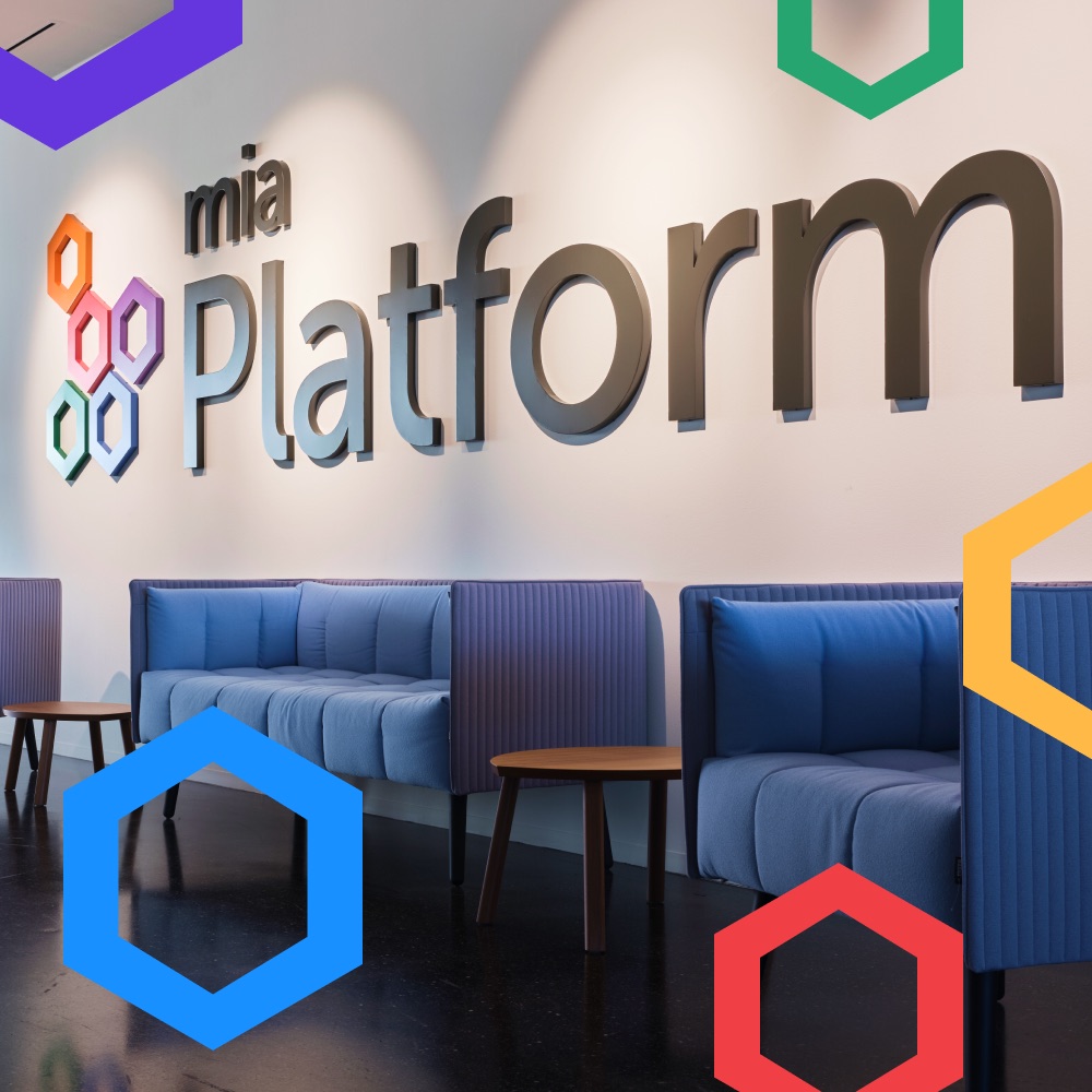 Mia‑Platform reception wall with the 3D company logo and lettering, blue lounge sofas, and colored hexagon outlines overlaid on the photo
